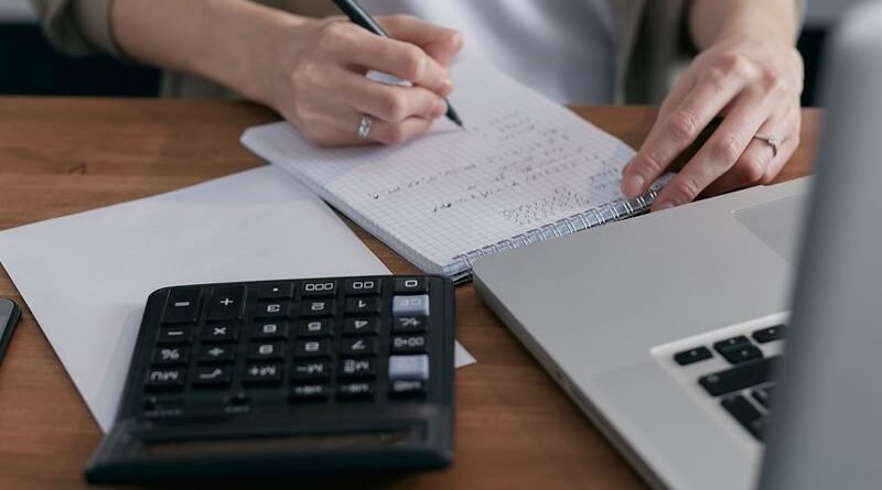 A woman writing financial calculations in a notebook with a calculator and laptop on a wooden desk