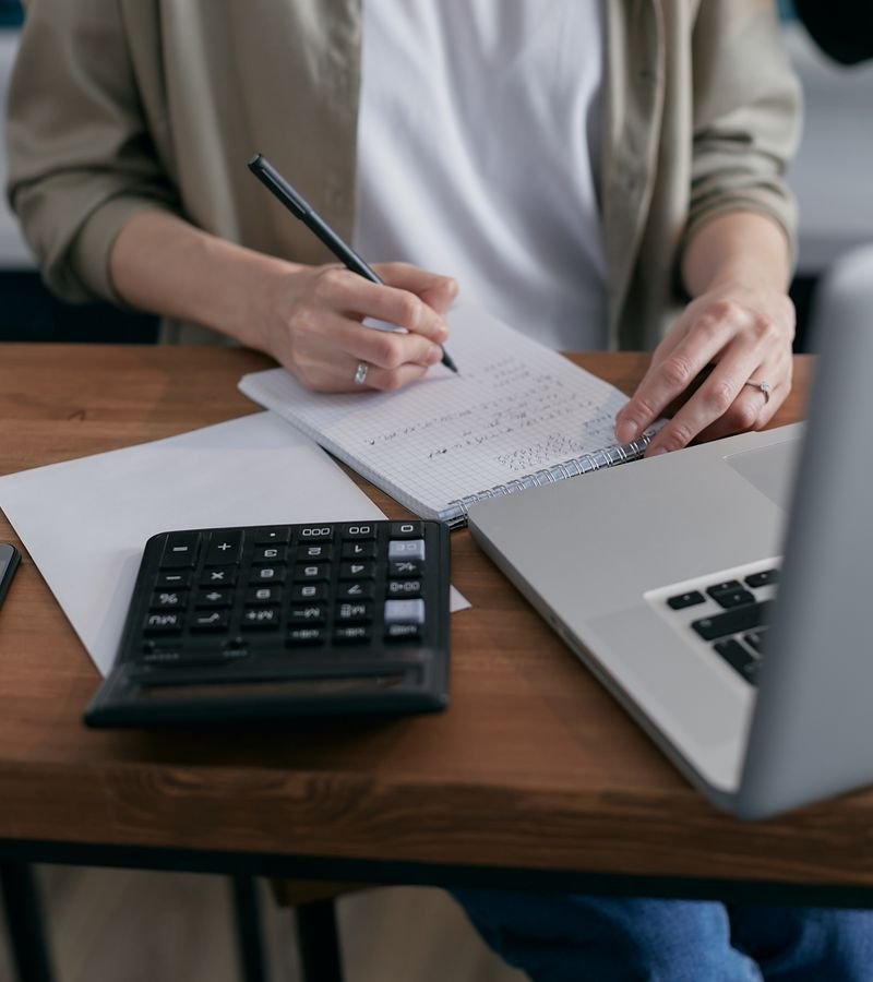 A woman writing financial calculations in a notebook with a calculator and laptop on a wooden desk
