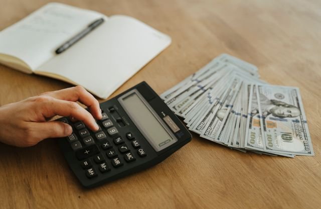 Close up of a hand using a calculator next to cash and a notebook on a wooden table