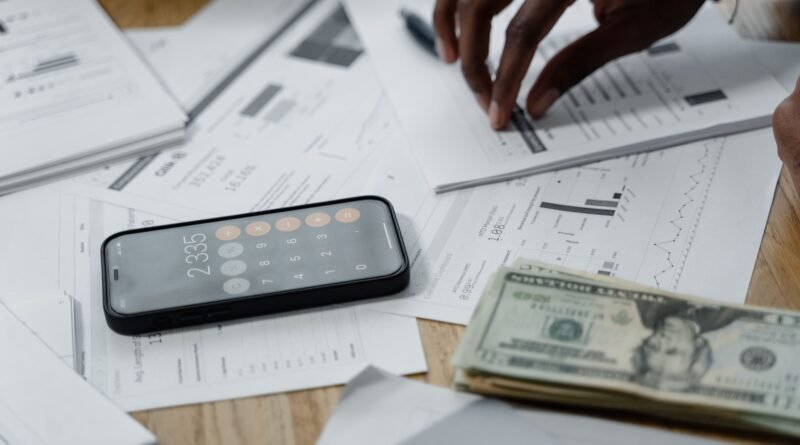 Close-up of hands calculating financial data with a smartphone, cash, and reports on a table