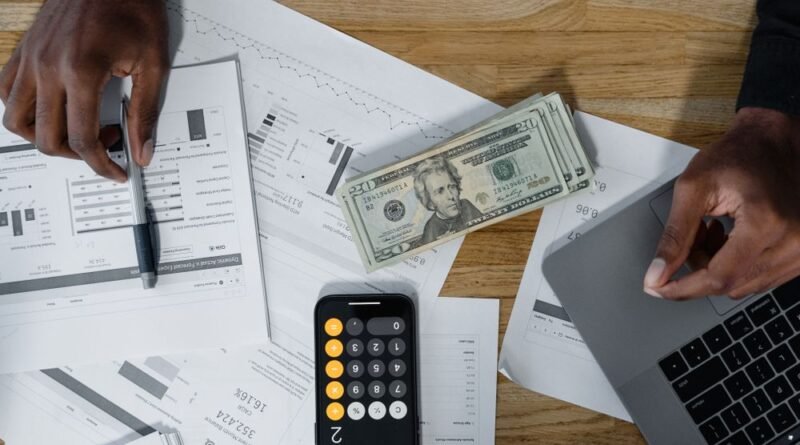 Hands calculating finances with papers, cash, and a laptop on a wooden desk