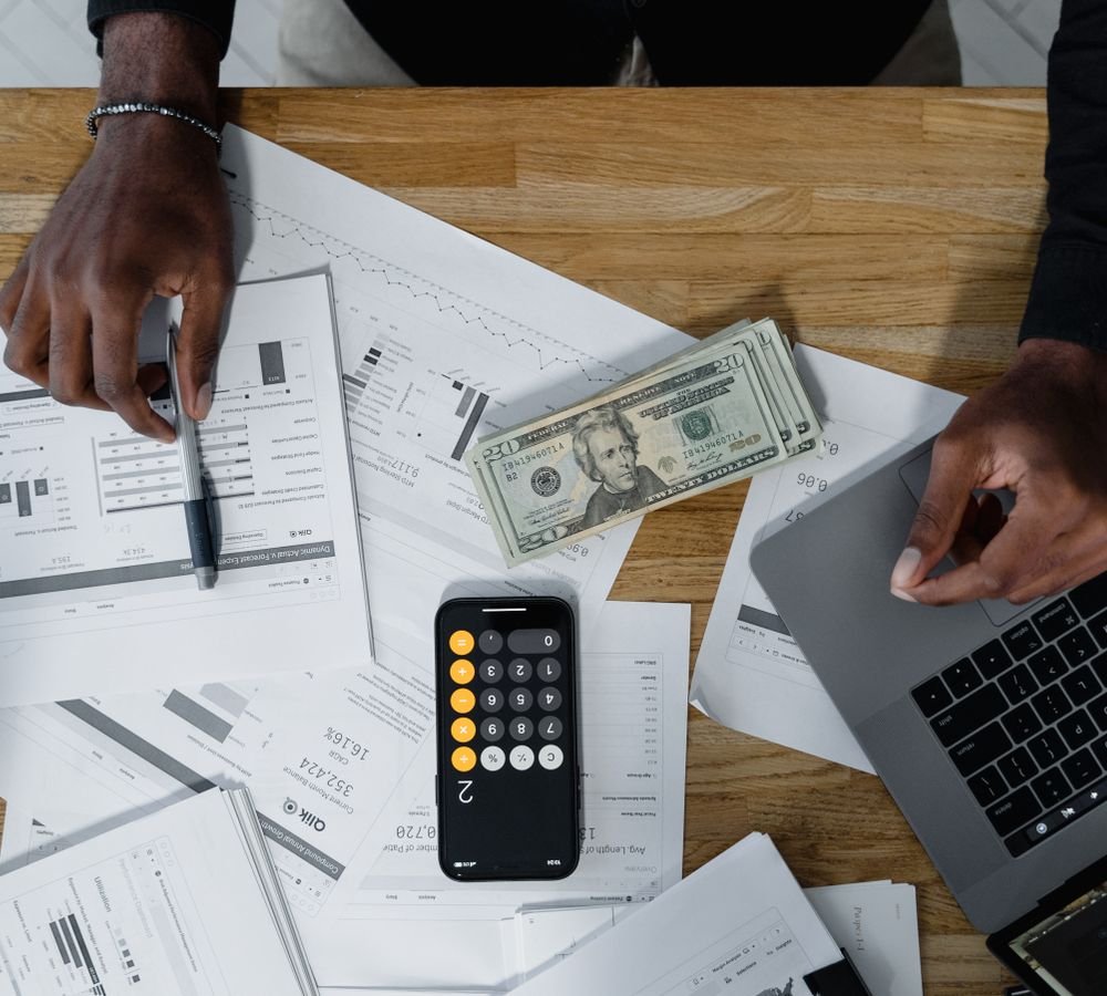 Hands calculating finances with papers, cash, and a laptop on a wooden desk