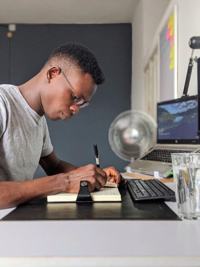 Young man writing in a notebook while working from home with a laptop and keyboard